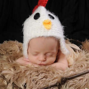 May include: A newborn baby wearing a white crocheted chicken hat with red, black, and yellow accents, sleeping in a brown faux fur basket.