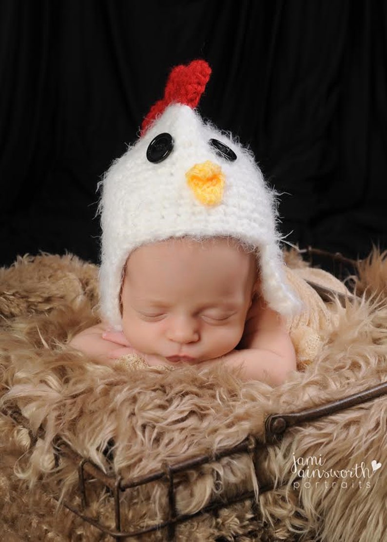 May include: A newborn baby wearing a white crocheted chicken hat with red, black, and yellow accents, sleeping in a brown faux fur basket.
