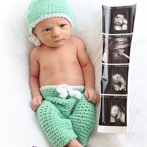 May include: A newborn infant wearing a mint green crocheted hat with a white trim and matching mint green crocheted pants with a white drawstring waist. Beside the infant is a vertical strip of four black and white ultrasound images.