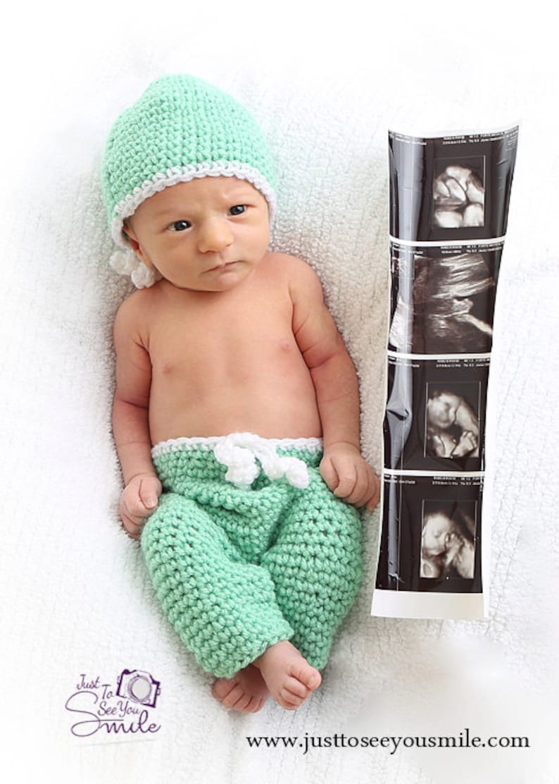 May include: A newborn infant wearing a mint green crocheted hat with a white trim and matching mint green crocheted pants with a white drawstring waist. Beside the infant is a vertical strip of four black and white ultrasound images.