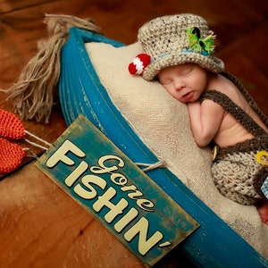 May include: A newborn baby wearing a brown and white crocheted outfit and a brown crocheted hat with a red and white fishing bobber on top. The baby is sleeping in a blue wooden boat with a sign that says "Gone Fishin'" on it. There are two orange crocheted fish on the wooden surface.