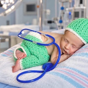 May include: A newborn infant sleeping while wearing a light green crocheted hat and matching green pants with white trim. A blue medical stethoscope is draped over the infant's body, resting on the white and blue striped fabric surface.