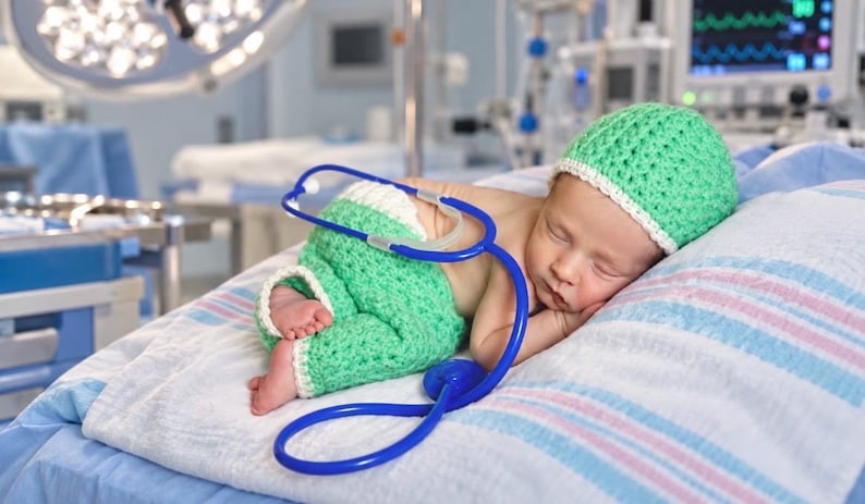May include: A newborn infant sleeping while wearing a light green crocheted hat and matching green pants with white trim. A blue medical stethoscope is draped over the infant's body, resting on the white and blue striped fabric surface.