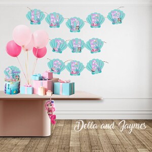 May include: A party decoration set featuring a banner with teal seashell-shaped cutouts displaying the letters for UNDER THE SEA, a blue cake with a seashell topper, pink balloons, wrapped gift boxes, and a small pink crocheted mermaid doll.