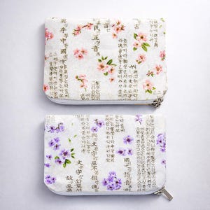 May include: Two rectangular, zippered pouches. The top pouch features pink floral patterns and text on a white background. The bottom pouch has purple floral patterns and text on a white background. Both pouches have silver zippers.