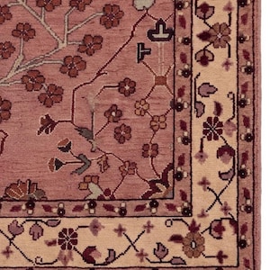 May include: A section of a detailed, hand-knotted rug. The rug features a dusty rose field with a floral pattern in shades of brown, burgundy, and cream. The border is cream with a repeating floral design. The rug has a fringe.