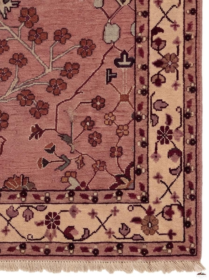 May include: A section of a detailed, hand-knotted rug. The rug features a dusty rose field with a floral pattern in shades of brown, burgundy, and cream. The border is cream with a repeating floral design. The rug has a fringe.