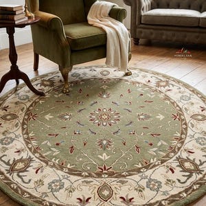May include: A circular rug with a floral pattern. The rug has a sage green central area and a cream-coloured border. The border is decorated with floral and leaf designs in brown, red, and blue. It is placed on a wooden floor.