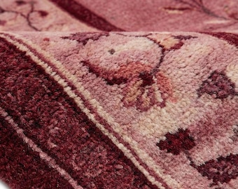 May include: Close-up of a section of a rug with a floral pattern. The rug features a deep burgundy border and a central field in shades of pink and burgundy. The floral design includes stylized flowers and leaves. The rug's texture is visible, suggesting a woven construction.