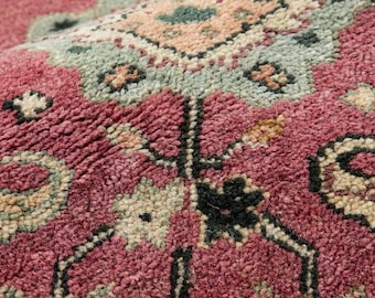 May include: Close-up of a rolled-up, intricately patterned rug. The rug features a deep rose-colored background with detailed floral and geometric designs in shades of green, beige, and black. The texture appears plush and woven, showcasing a traditional style.