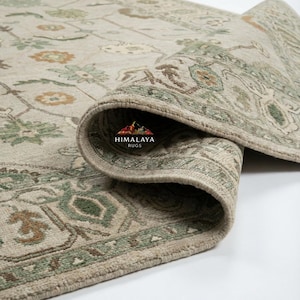 May include: Beige and green area rug with a rolled edge, showcasing a detailed floral pattern. The rug features a border design and a logo that reads "Himalaya Rugs".