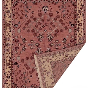 May include: A rectangular, hand-woven rug with a floral design in shades of rose, cream, and burgundy. The rug features a central field with a repeating floral pattern and a cream-colored border with intricate floral motifs. The corner is folded back, revealing the underside.