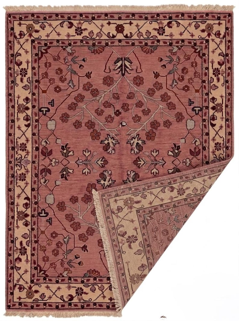 May include: A rectangular, hand-woven rug with a floral design in shades of rose, cream, and burgundy. The rug features a central field with a repeating floral pattern and a cream-colored border with intricate floral motifs. The corner is folded back, revealing the underside.