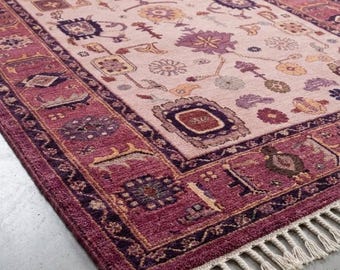 May include: A rectangular area rug featuring a light pink center field with scattered floral and geometric motifs in purple, orange, and dark red. The rug has a wide, dark magenta border with intricate patterns and off-white fringe on the ends.
