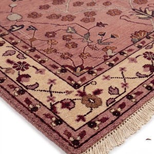 May include: A detailed close-up of a rug with a floral pattern. The rug features a dusty rose field with a cream-colored border. The border is adorned with floral and geometric designs in shades of burgundy and brown. The rug has a fringe.