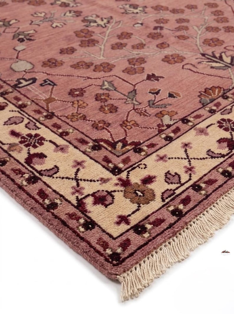 May include: A detailed close-up of a rug with a floral pattern. The rug features a dusty rose field with a cream-colored border. The border is adorned with floral and geometric designs in shades of burgundy and brown. The rug has a fringe.