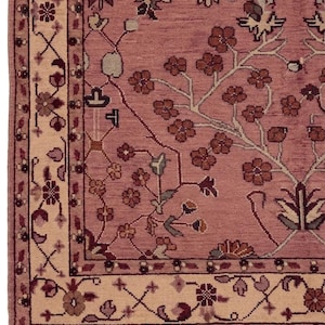 May include: Close-up of a dusty rose-colored rug with a floral pattern. The rug features a cream-colored border with intricate floral and geometric designs. The design includes brown, burgundy, and green accents. The rug has a fringed edge.