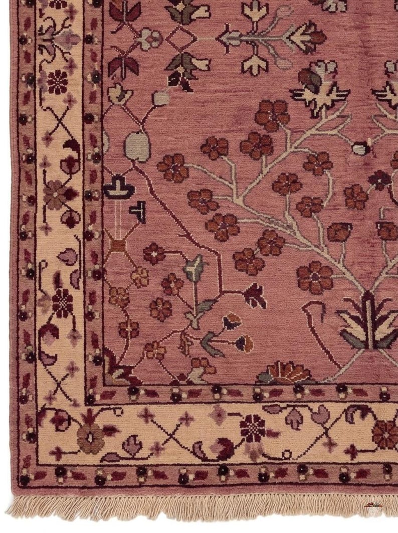 May include: Close-up of a dusty rose-colored rug with a floral pattern. The rug features a cream-colored border with intricate floral and geometric designs. The design includes brown, burgundy, and green accents. The rug has a fringed edge.