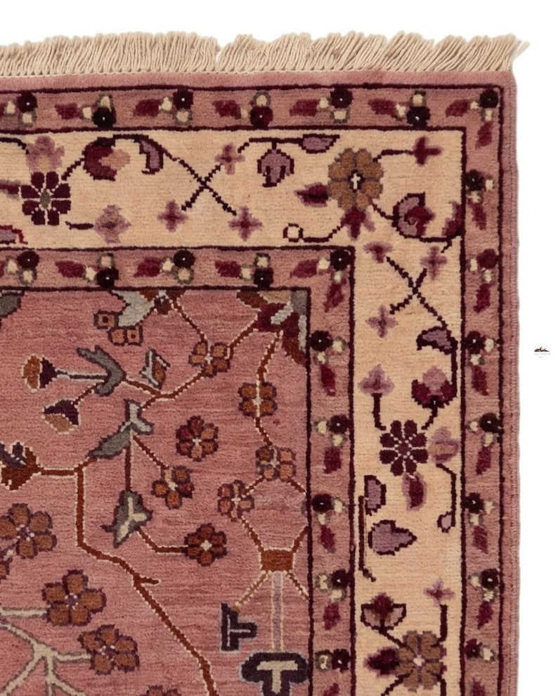 May include: Close-up of a section of a rug with a floral design. The rug features a dusty rose field with a cream-colored border. The border and field are adorned with floral and vine patterns in shades of burgundy, brown, and green.