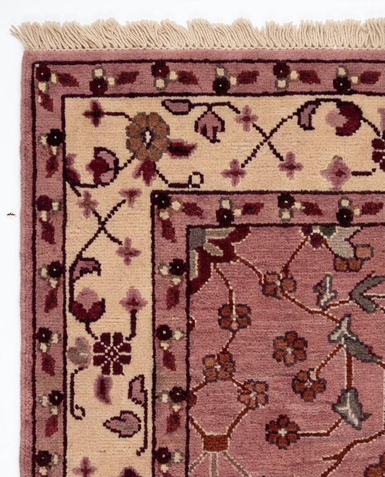 May include: Close-up of a section of a rectangular rug with a floral design. The rug features a light pink field with a cream-colored border and a darker pink border. The design includes floral and vine motifs in shades of pink, burgundy, and brown.