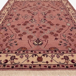 May include: A rectangular rug with a dusty rose field and a cream-colored border. The rug features a floral pattern with brown, burgundy, and green accents. The rug has a fringe on one end.