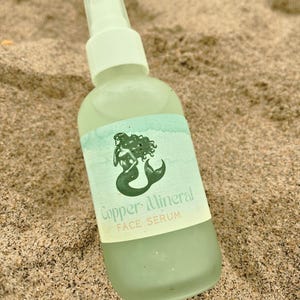 May include: A light green glass bottle of Copper Mineral Face Serum with a white pump top. The label features a green mermaid illustration. The bottle is resting on sand.