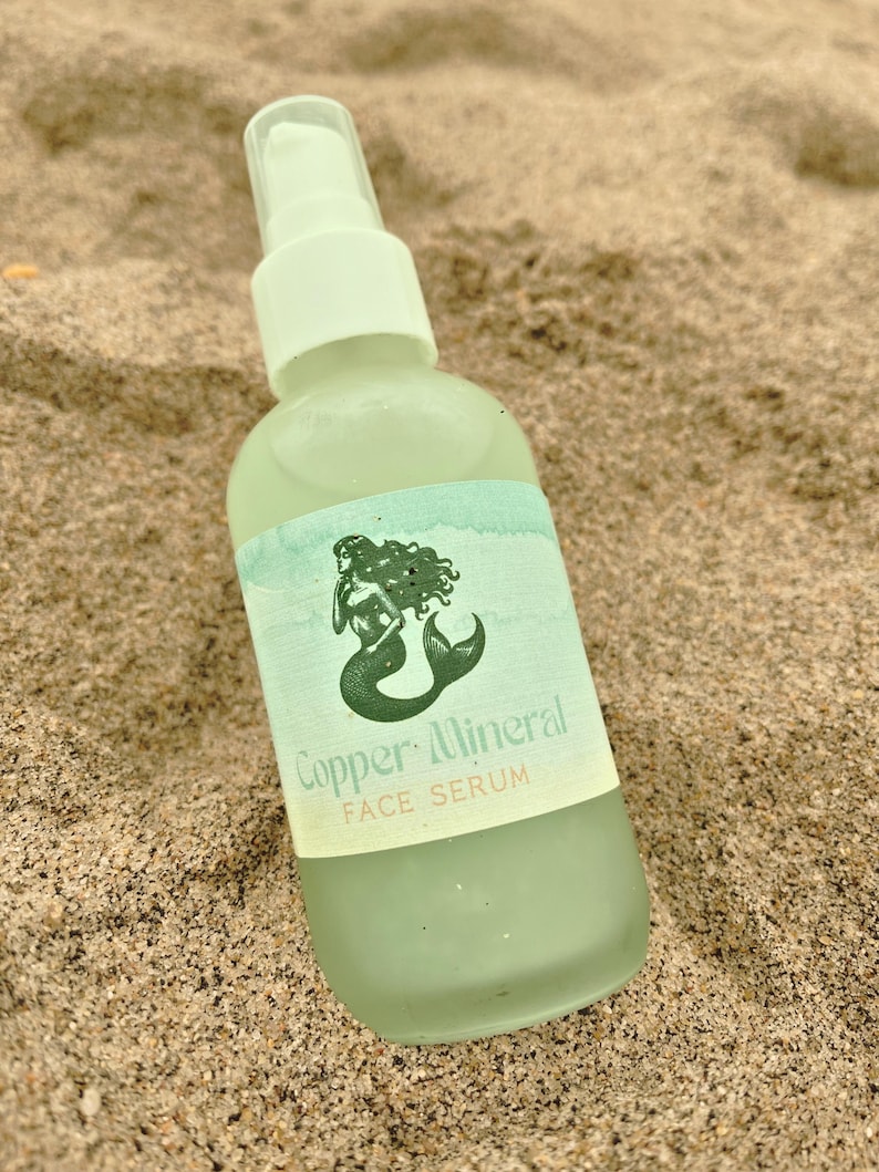 May include: A light green glass bottle of Copper Mineral Face Serum with a white pump top. The label features a green mermaid illustration. The bottle is resting on sand.