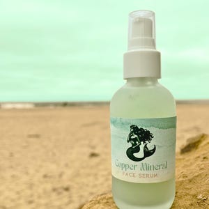 May include: A frosted glass bottle of Copper Mineral Face Serum with a white pump top. The label features a black and white illustration of a mermaid. The background shows a beach with a light blue sky.