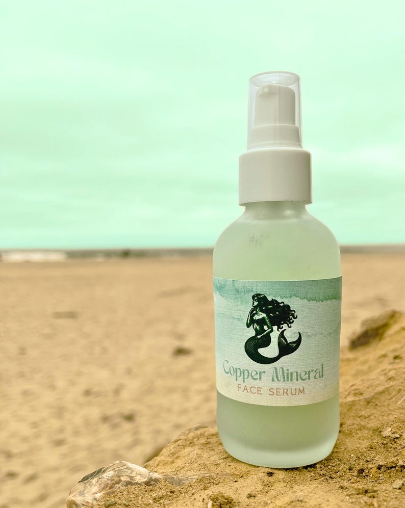 May include: A frosted glass bottle of Copper Mineral Face Serum with a white pump top. The label features a black and white illustration of a mermaid. The background shows a beach with a light blue sky.