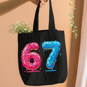 May include: Black tote bag with the numbers 6 and 7 designed as donuts. The number 6 is pink with sprinkles, and the number 7 is blue with sprinkles. The bag has black handles and is filled with white flowers.