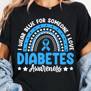 May include: A black t-shirt featuring a graphic design with a blue rainbow and a light blue awareness ribbon in the center. The text reads I WEAR BLUE FOR SOMEONE I LOVE DIABETES Awareness, with white stars on either side of the word Awareness.