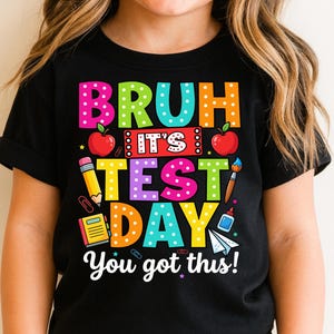 May include: Black t-shirt with a colorful graphic that reads "BRUH IT'S TEST DAY You got this!" The design includes apples, a pencil, a paintbrush, and other school-themed elements. The text is in various bright colors.