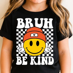 May include: Black t-shirt featuring a yellow smiley face wearing an orange beanie and making a peace sign. The shirt has the words "BRUH" and "BE KIND" in white block letters, set against a black and white checkered background.