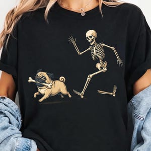 Könnte beinhalten: Ein schwarzes T-Shirt mit einem Grafikdruck eines beigen Mopses, der mit einem Knochen im Maul rennt, gefolgt von einem ebenfalls rennenden menschlichen Skelett. Dem Skelett scheint der Unterschenkelknochen zu fehlen, der separat abgebildet ist.