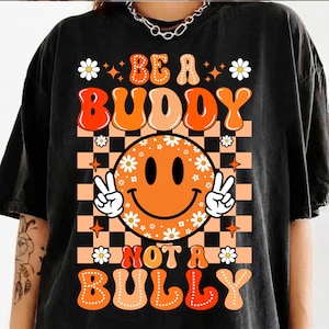 Könnte beinhalten: Ein schwarzes T-Shirt mit einem Grafikdruck und dem Text BE A BUDDY NOT A BULLY. Das Design zeigt ein orangefarbenes Smiley-Gesicht mit weißen Gänseblümchen, einen pfirsichfarbenen und schwarzen Karohintergrund sowie zwei Hände mit Friedenszeichen.