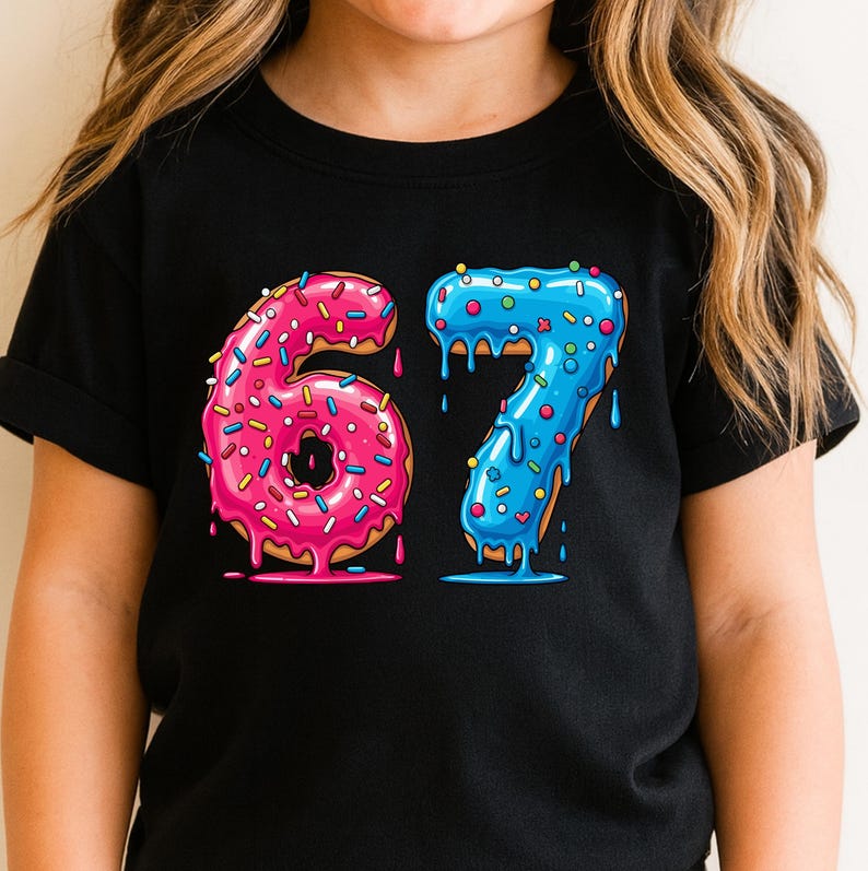 May include: Black t-shirt with a graphic of the numbers "67" designed as donuts. The number "6" is pink with pink and blue sprinkles, and the number "7" is blue with blue sprinkles. The donuts have dripping icing.