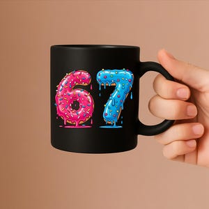 May include: Black ceramic mug with the numbers "67" designed as donuts. The number "6" is pink with sprinkles, and the number "7" is blue with sprinkles. The mug has a black handle. The mug is held by a hand.