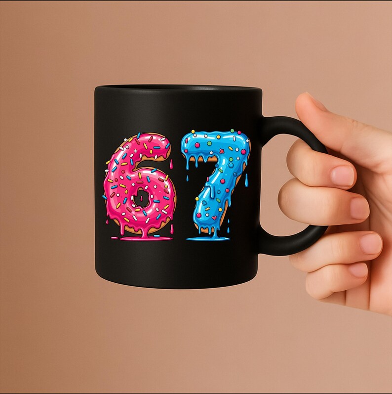 May include: Black ceramic mug with the numbers "67" designed as donuts. The number "6" is pink with sprinkles, and the number "7" is blue with sprinkles. The mug has a black handle. The mug is held by a hand.