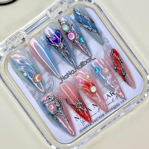 Puede incluir: Un conjunto de uñas postizas largas en forma de almendra en una caja de plástico transparente. Las uñas presentan una variedad de diseños, incluyendo tonos iridiscentes, azules, rosas y rojos, con detalles plateados y de joyas. El texto "NaNa.Nailbox" es visible.
