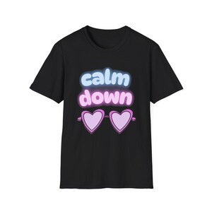 Puede incluir: Camiseta negra con las palabras "calm down" en una fuente de estilo neón, con colores azul y rosa. Debajo del texto hay un par de gafas de sol rosas en forma de corazón. Prenda de vestir informal.