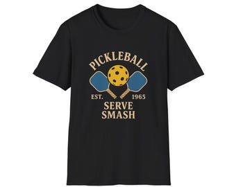 Może przedstawiać: Czarna koszulka z nadrukiem do gry w pickleball. Projekt zawiera żółtą piłkę do pickleball, dwie niebieskie rakiety oraz napisy "PICKLEBALL", "EST. 1965" i "SERVE SMASH" w stylu vintage. Koszulka wykonana jest z miękkiego materiału.