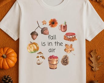 Puede incluir: Camiseta blanca con un diseño de acuarela con elementos otoñales. El diseño incluye bellotas, una calabaza, setas, un girasol, una taza, un cubo de manzanas, una piña y hojas. El texto dice "fall is in the air."