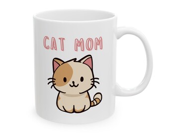 Könnte beinhalten: Weiße Keramiktasse mit dem Text "CAT MOM" in rosa Buchstaben. Die Tasse zeigt eine Cartoon-Katzenillustration mit tan- und braunfarbenem Fell. Die Katze hat ein lächelndes Gesicht und einen gestreiften Schwanz.