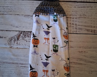 May include: A hanging kitchen towel featuring a white fabric with a Halloween-themed print of cocktails, pumpkins, ghosts, and bats. The top section is made of grey crocheted yarn and includes a single light-colored wooden button for hanging.