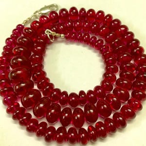 Pode incluir: Um colar composto por vários fios de contas polidas, vermelho escuro. As contas variam em tamanho, sendo as maiores na frente e as menores atrás. Um fecho prateado prende o colar.