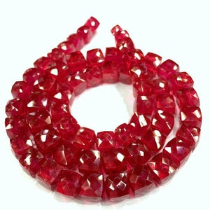 May include: A necklace of faceted, square-cut ruby beads. The beads are a deep red colour and appear to be strung together. The gemstones are displayed against a white background, highlighting their brilliance and clarity. The necklace is a jewellery item.