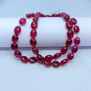 May include: Two strands of oval-shaped, deep red gemstones. The polished stones are strung together, showcasing their translucent quality. The beads are a rich, vibrant color, reflecting light and creating a luxurious appearance. The gemstones are likely used for jewelry making.