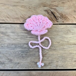 May include: A handmade, light pink crochet flower with a detailed petal design. The flower has a stem with a looped design and three small white beads at the end. The craft item is set against a wooden background.