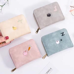 Pode incluir: Quatro carteiras pequenas e quadradas em creme, cinza, rosa e azul-esverdeado. Cada carteira tem um fecho de zíper e um design bordado exclusivo, incluindo um porco com uma cenoura, uma ovelha dormindo e um gato com um coração.
