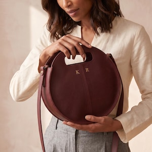 Può includere: Una borsa a mano in pelle bordeaux dal design circolare. La borsa presenta un manico superiore e una lunga tracolla. Le iniziali dorate "K R" sono impresse sul davanti. La borsa è tenuta da una persona che indossa una camicia color crema e pantaloni grigi.
